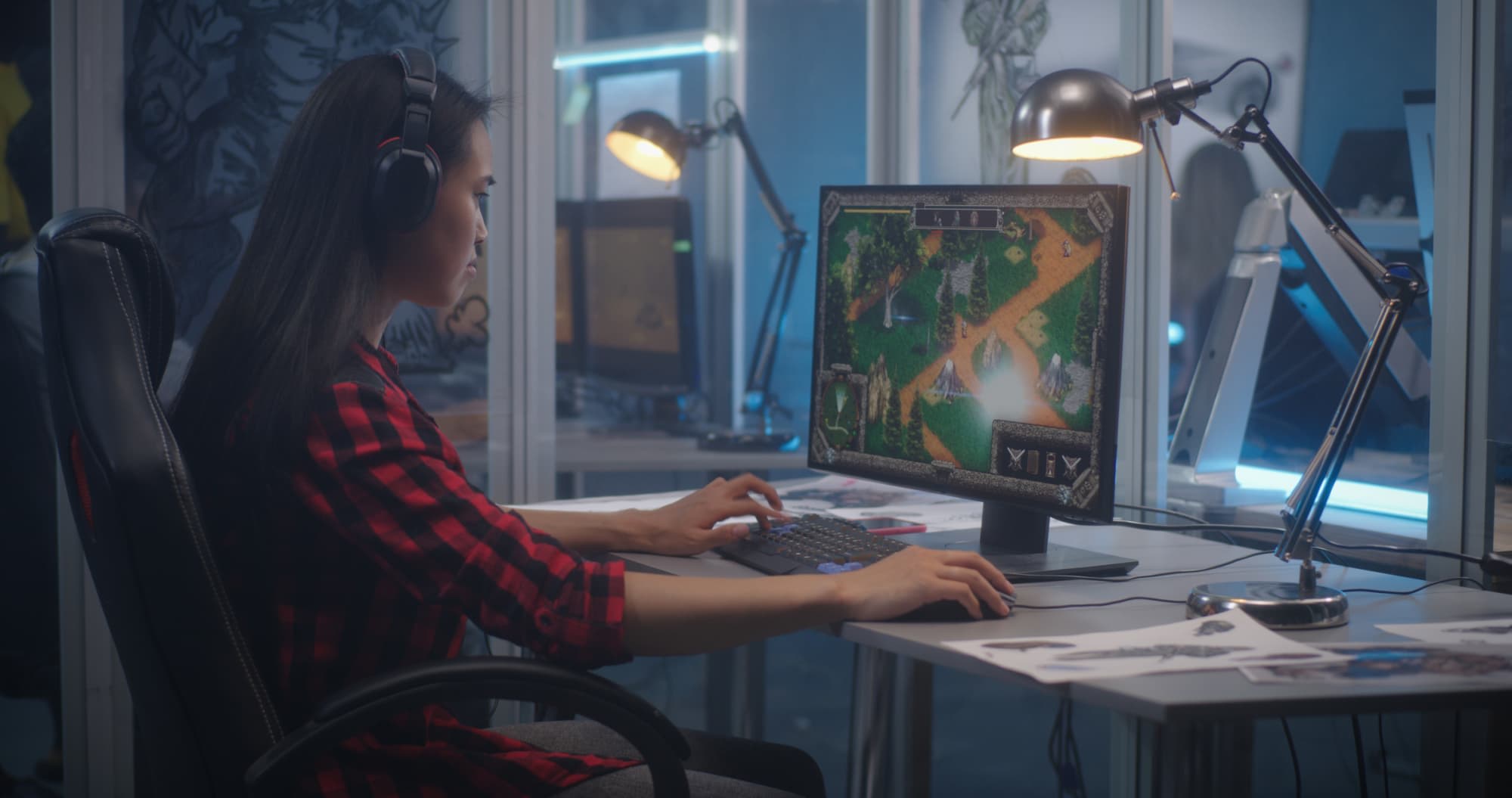 A female game developer wearing headphones works on a computer, developing a game in an office setting.